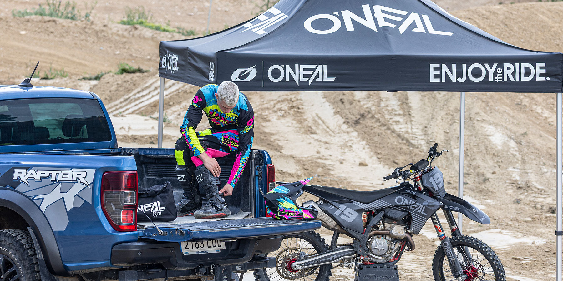 Ein Motocross-Fahrer in farbenfroher Ausrüstung sitzt auf der offenen Heckklappe eines blauen Ford Raptor Trucks und stellt seine Stiefel neben einem schwarzen Dirt Bike und unter einem schwarzen O'Neal-Vordach auf einer Dirt Track im Freien ein.