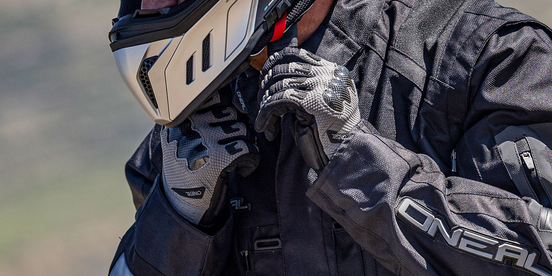 Gros plan sur une personne portant une veste de moto noire, des gants d'équitation et un casque, et fixant la sangle du casque. Le nom de la marque "O'NEAL" est visible sur la veste et les gants.
