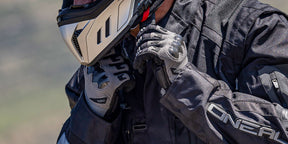 Primo piano di una persona che indossa una giacca da moto nera, guanti da equitazione e un casco e allaccia il cinturino del casco. Il marchio "O'NEAL" è visibile sulla giacca e sui guanti.