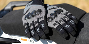 Un paio di guanti da moto neri e grigi con protezioni per le nocche sono appoggiati su un sedile nero. Una scritta parziale "READY TO RACE" è visibile su una superficie bianca nelle vicinanze.