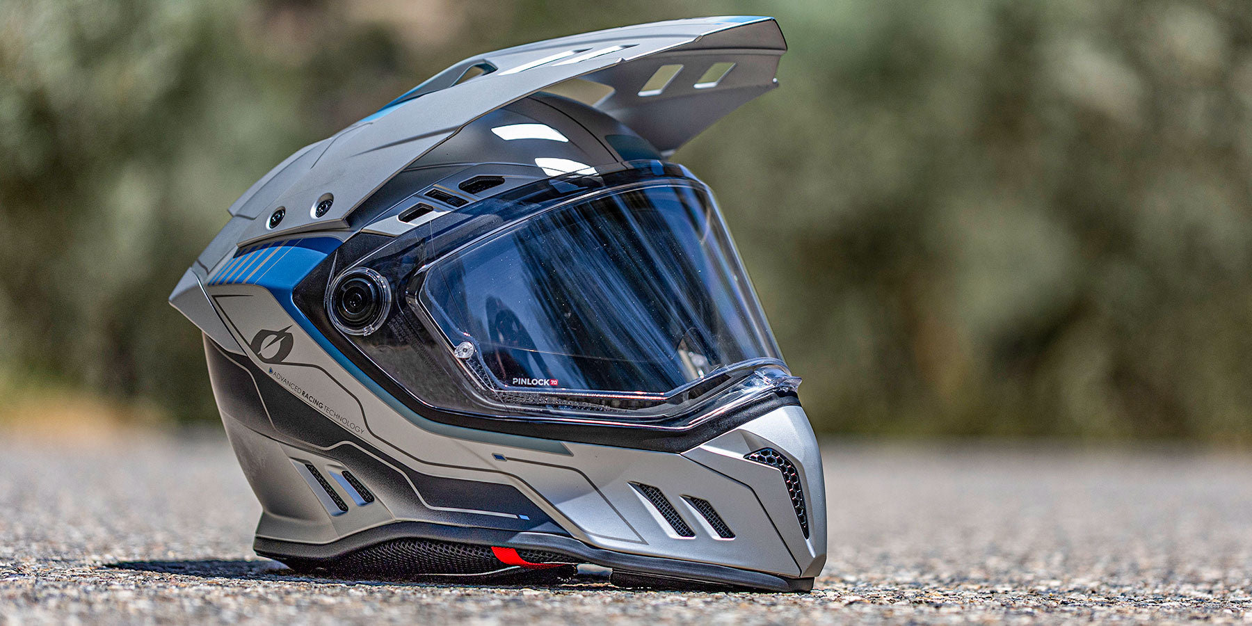 Ein silberner und blauer Offroad-Motorradhelm mit klarem Visier steht auf einer Asphaltfläche mit einem unscharfen natürlichen Hintergrund.