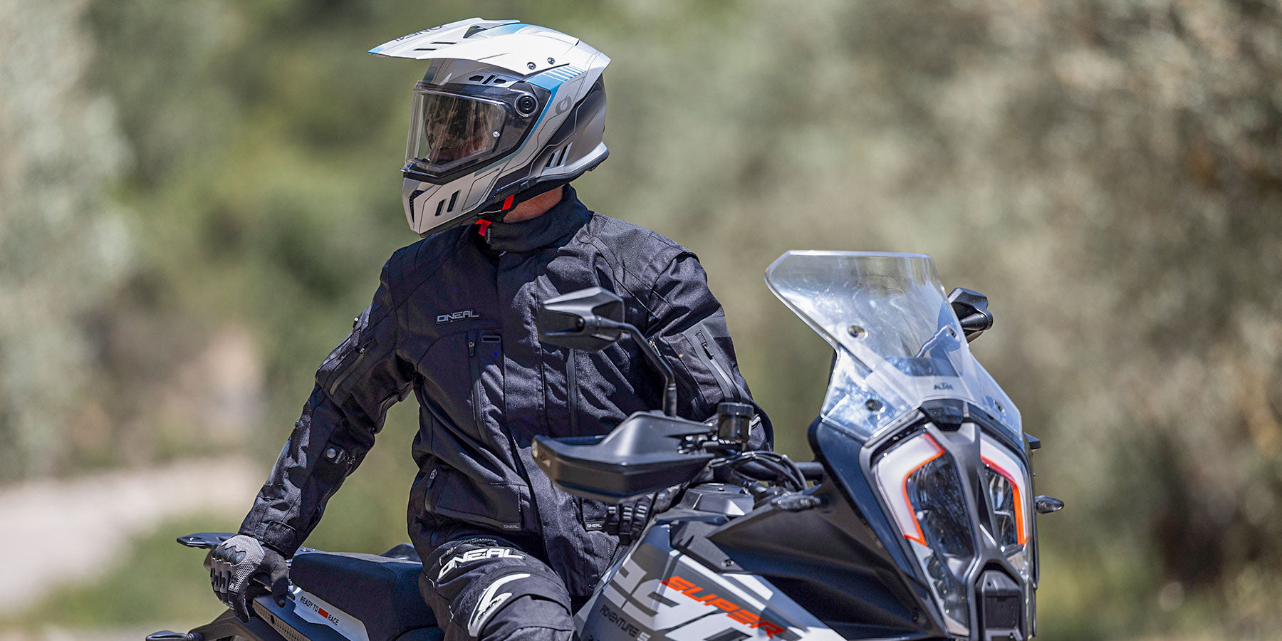 Eine Person, die eine schwarze Motorradjacke und einen weißen Helm trägt, sitzt auf einem Motorrad im Freien, wobei die Bäume im Hintergrund verschwimmen.