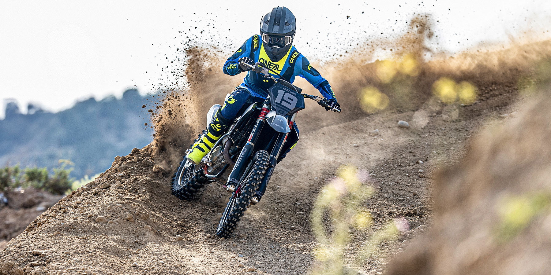 Ein Motocross-Fahrer in blauer und gelber Ausrüstung rast um eine Kurve auf einer unbefestigten Strecke und wirbelt dabei Staub und Schmutz auf, während im Hintergrund felsiges Gelände und unscharfes Laub zu sehen sind.