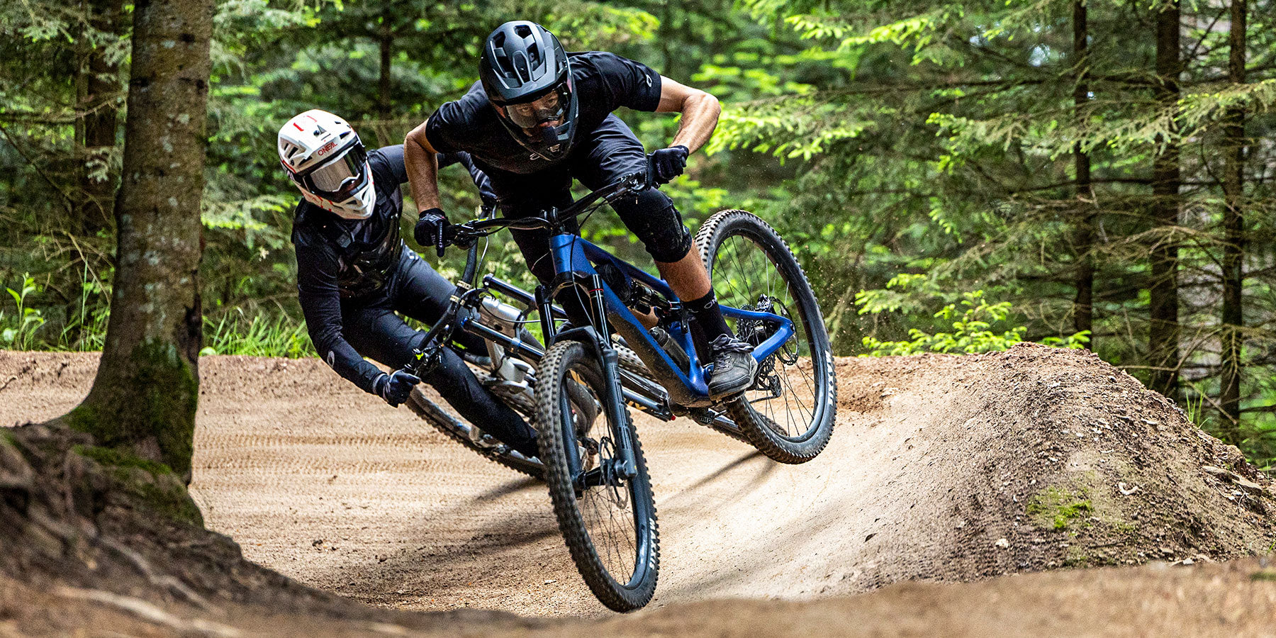 Zwei Mountainbiker mit Helmen und Schutzausrüstung lehnen sich in eine Kurve eines unbefestigten Waldweges und schleudern Schmutz auf, während sie den von Bäumen gesäumten Pfad entlang rasen.