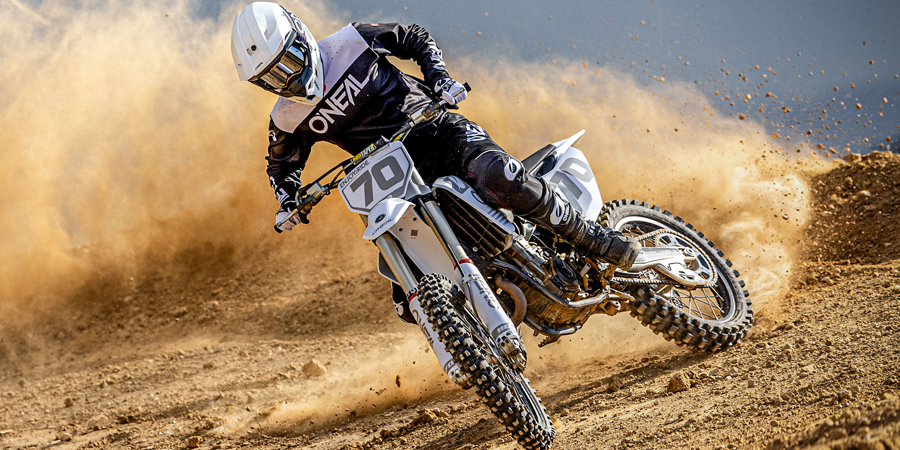 Ein Motocross-Fahrer in schwarzer und weißer Kleidung biegt auf einer unbefestigten Strecke in eine Kurve ein und wirbelt dabei eine große Staubwolke auf. Der Fahrer trägt zum Schutz einen Helm und eine Schutzbrille.