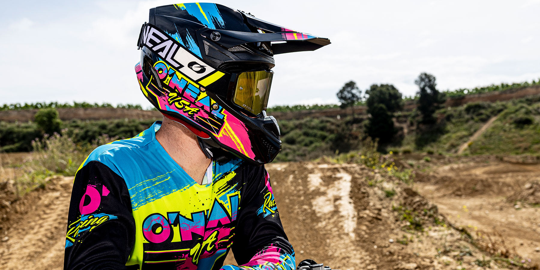 Ein Motocross-Fahrer mit buntem Helm und Trikot blickt zur Seite auf einer unbefestigten Strecke mit Grün und Bäumen im Hintergrund unter einem strahlenden Himmel.