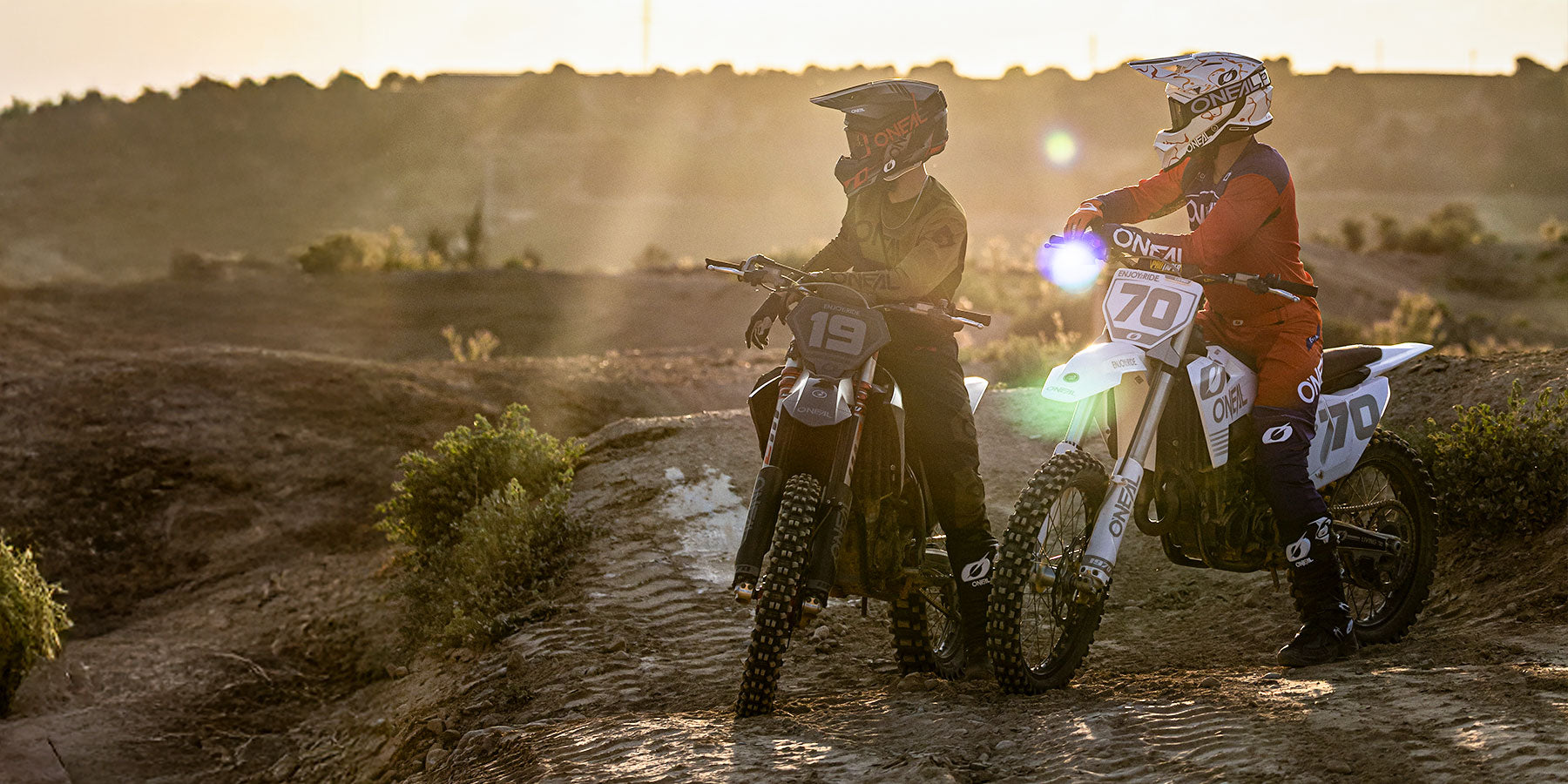 Zwei Motocross-Fahrer mit Helmen und Ausrüstung sitzen auf Dirt-Bikes auf einer unbefestigten Strecke bei Sonnenuntergang, umgeben von rauem Gelände und sanftem Sonnenlicht.
