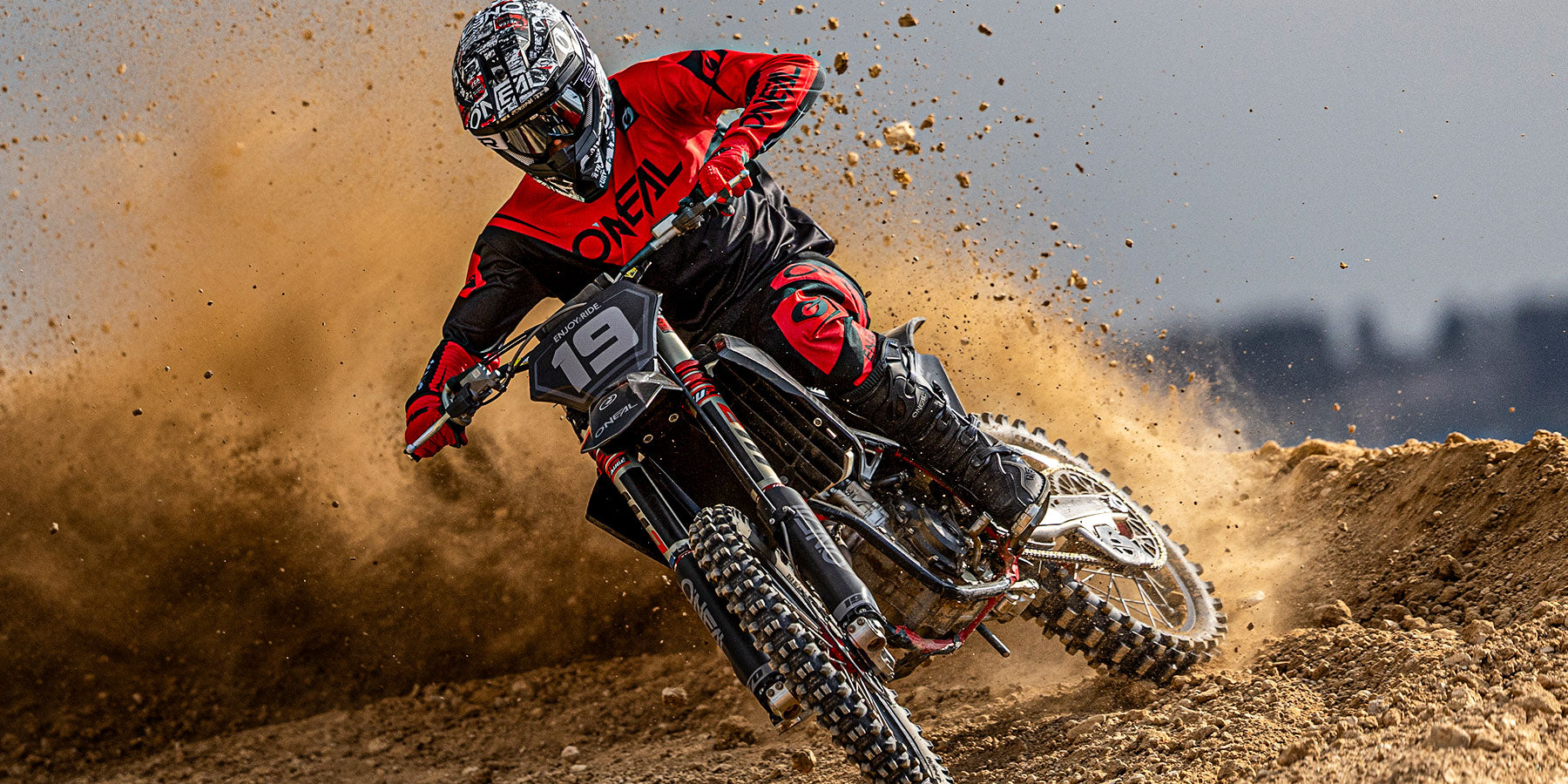 Ein rot-schwarz gekleideter Motocross-Fahrer rast um eine Kurve einer Dirt-Track-Strecke und wirbelt dabei eine Staubwolke auf, während er den Lenker eines nummerierten Dirt-Bikes umklammert.