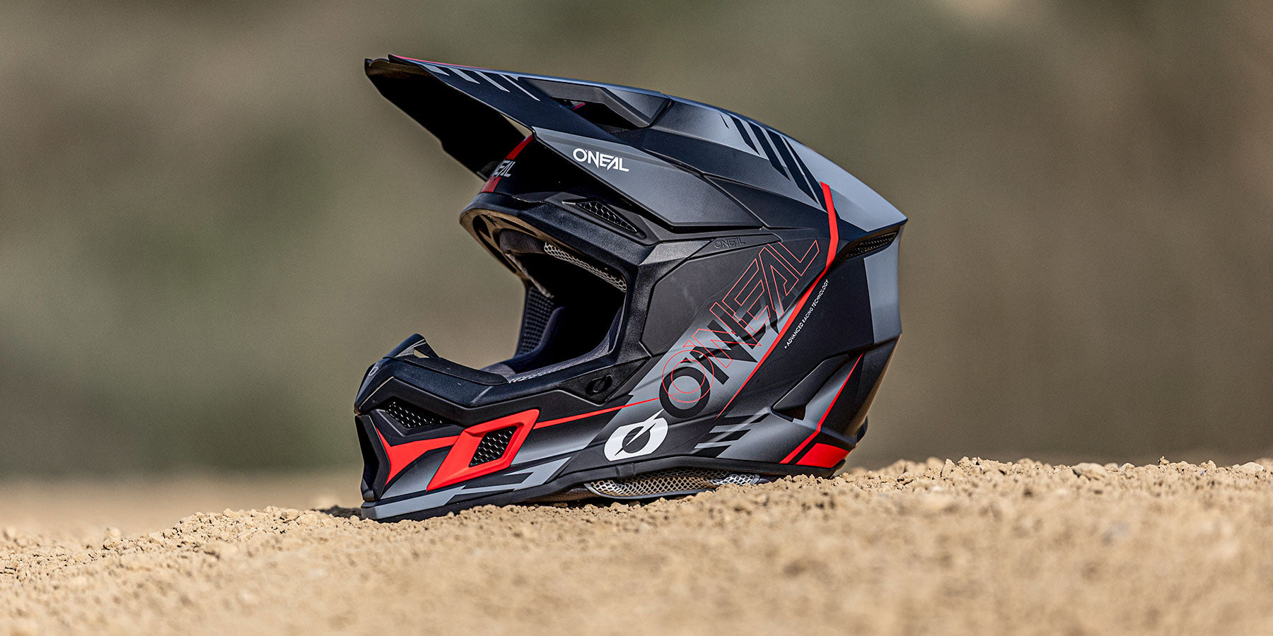 Ein schwarzer und roter O'Neal-Motocross-Helm liegt im Freien auf sandigem Boden, der Hintergrund ist unscharf.