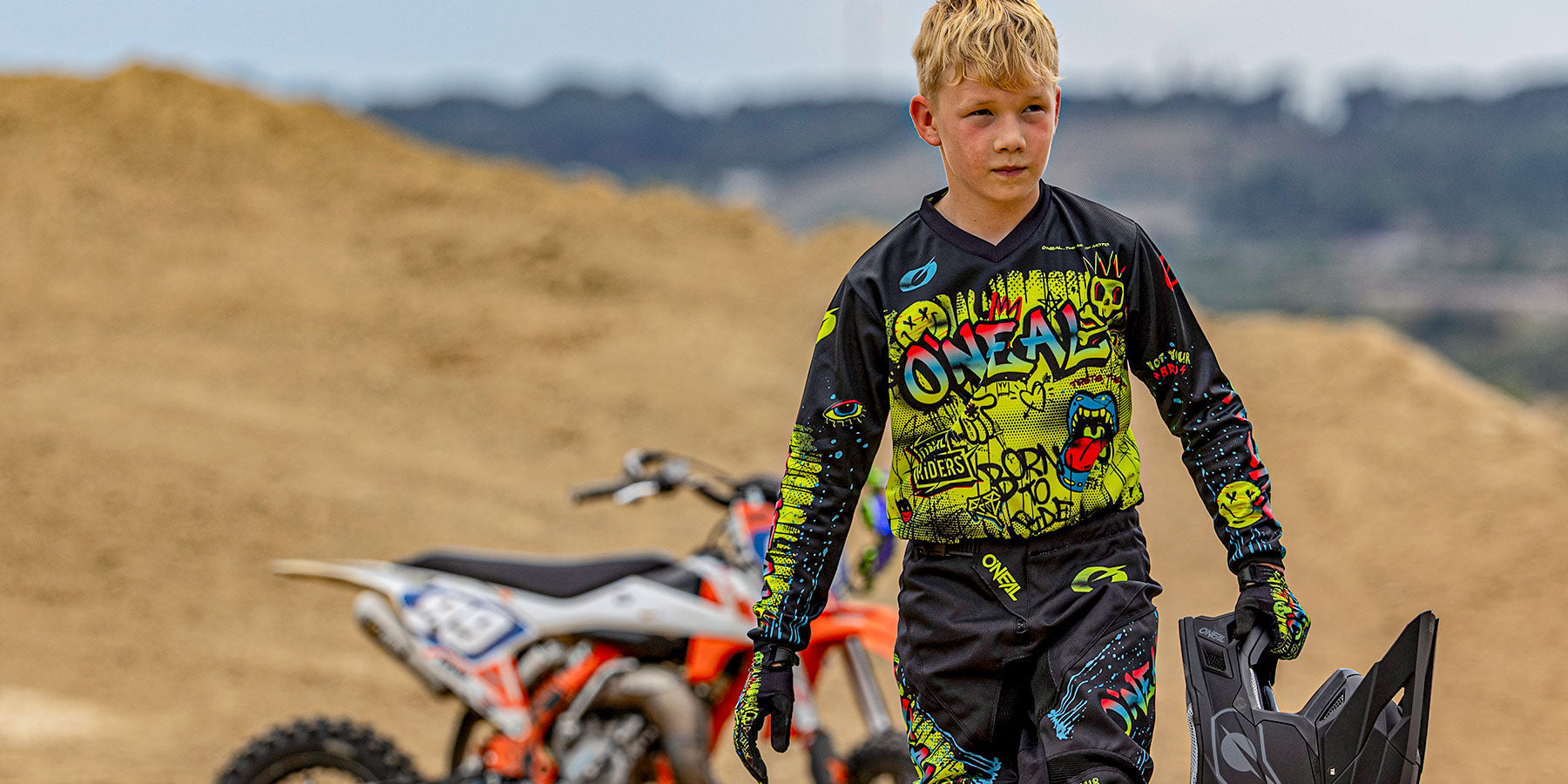Ein Junge in farbenfroher Motocross-Ausrüstung trägt einen Helm und läuft auf einer unbefestigten Strecke, mit zwei Motorrädern und sandigen Hügeln im Hintergrund.