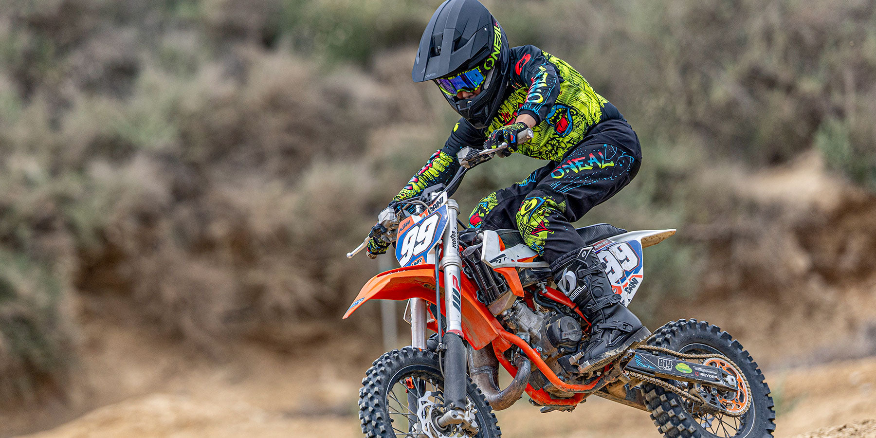 Ein Motocross-Fahrer in schwarzer und neongrüner Kleidung und mit einem Helm fährt auf einem orangefarbenen Dirt Bike mit der Nummer 39 über eine unbefestigte Strecke.