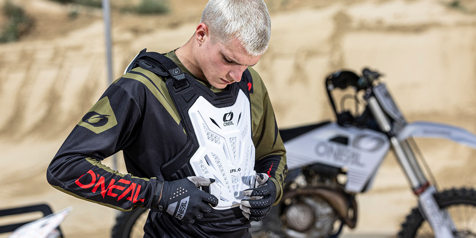 Ein junger Motocross-Fahrer in Schutzkleidung zieht seinen weißen Brustpanzer an. Er befindet sich im Freien in der Nähe eines Dirt-Bikes mit sandigem Gelände im Hintergrund.