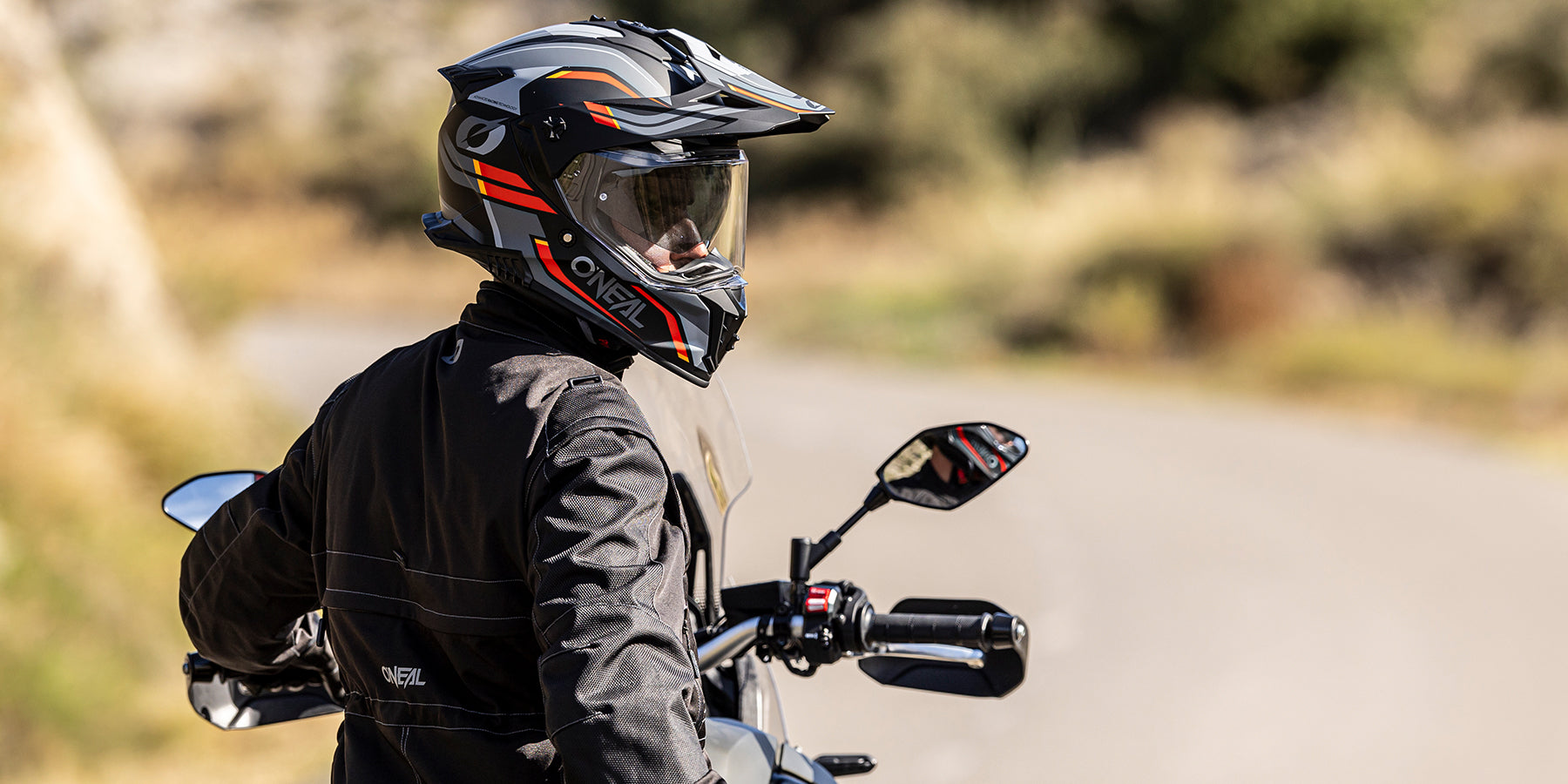 Eine Person, die einen schwarzen O'Neal-Motorradhelm und eine Jacke trägt, sitzt auf einem Motorrad und blickt auf eine kurvenreiche Straße im Freien mit unscharfen Bäumen und Gras im Hintergrund.