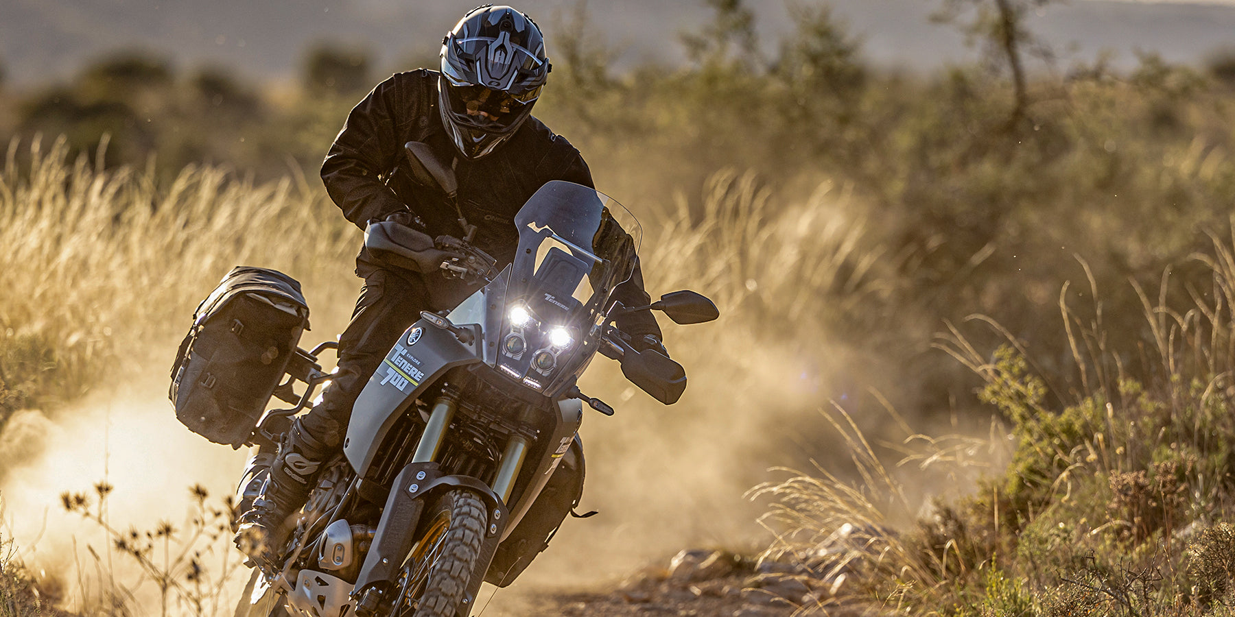 Eine Person, die einen Helm und eine Schutzausrüstung trägt, fährt mit einem Adventure-Motorrad auf einer staubigen, zerklüfteten Strecke, umgeben von hohem Gras und Sonnenlicht.