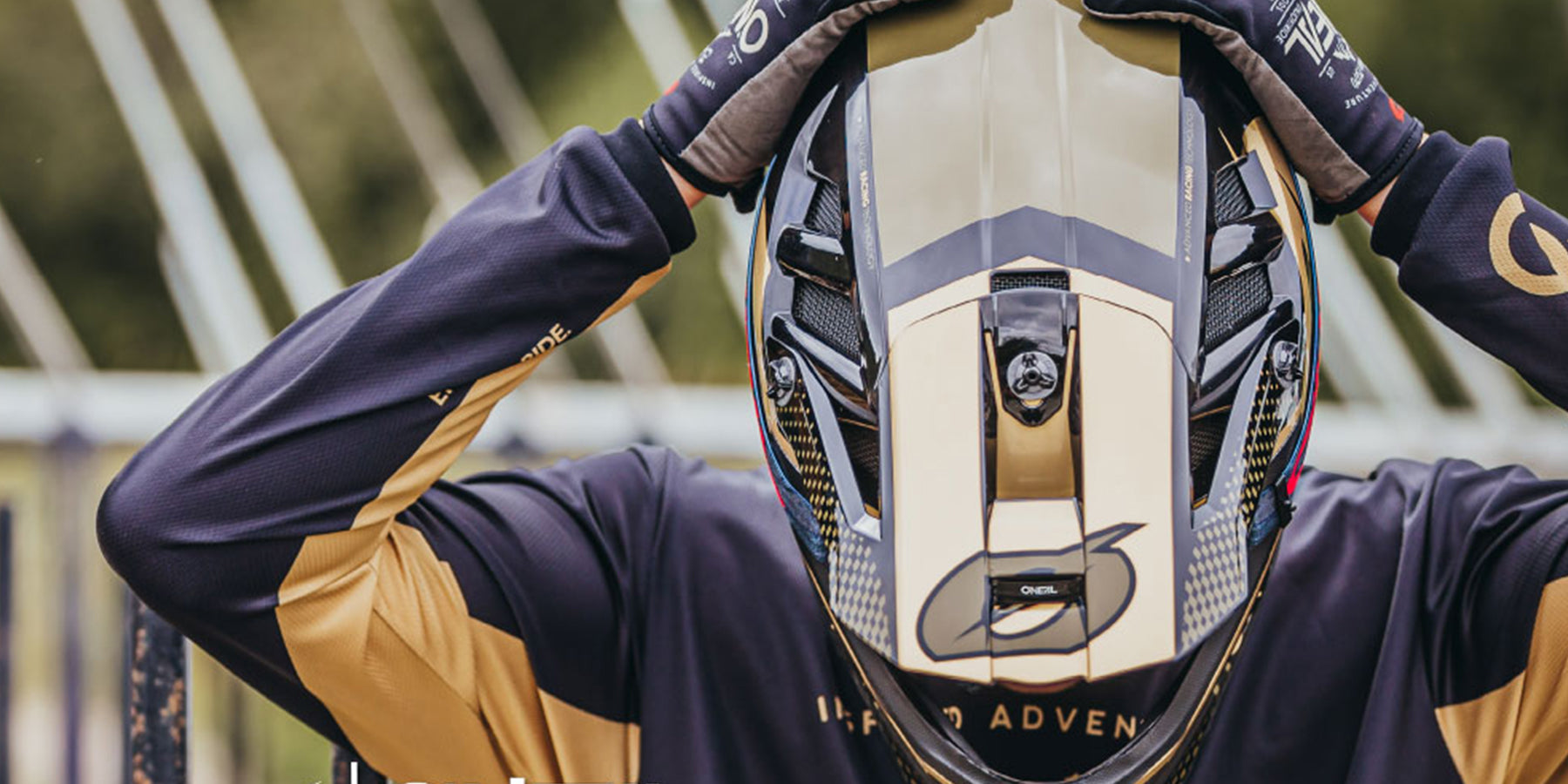 Person mit einem schwarz-goldenen Motorradhelm und passenden Handschuhen, die Arme sind so ausgestreckt, dass sie die Seiten des Helms berühren. Der Hintergrund ist verschwommen und weist Grüntöne auf, die eine Umgebung im Freien suggerieren.