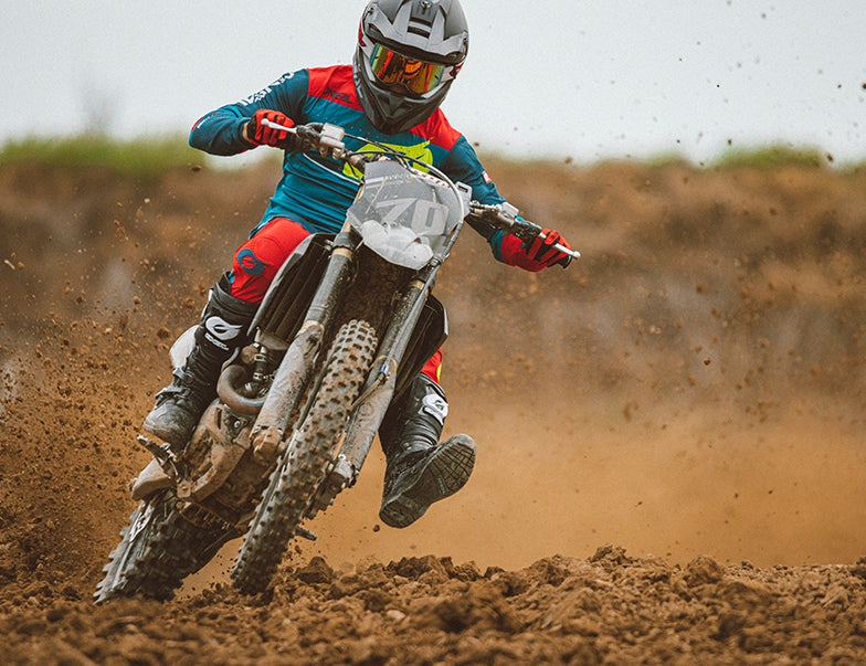 Ein Motocrossfahrer in voller Montur legt sich in eine Kurve und wirbelt dabei Staub auf, während er auf einer holprigen Strecke rast. Der Fahrer trägt einen Helm mit getöntem Visier und einen bunten Anzug. Der Hintergrund ist eine verschwommene Landschaft aus braunem Staub und spärlichem Grün.