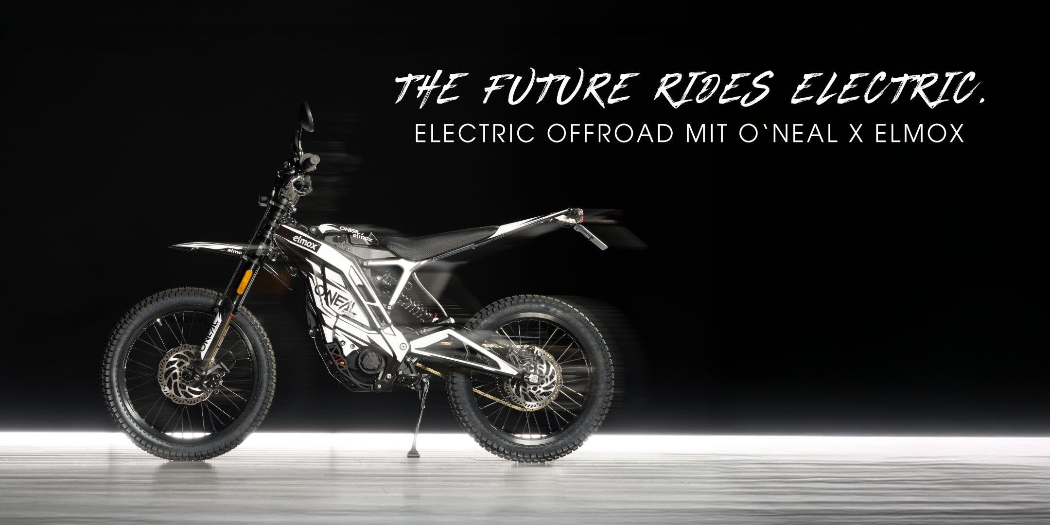 Ein schnittiges schwarz-weißes Elektro-Offroad-Motorrad ist auf einer glatten Oberfläche abgebildet, darüber steht der Text "The Future Rides Electric. Electric Offroad mit O'Neal x Elmox" steht in fetter, stilvoller Schrift darüber.