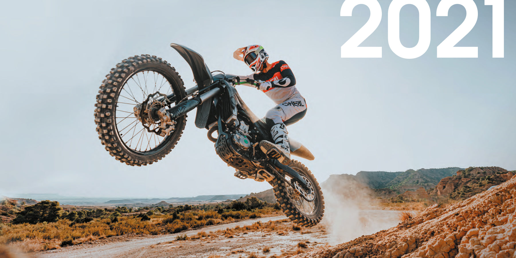 Ein Motocrossfahrer in voller Montur vollführt einen Sprung auf einem Dirtbike in einer rauen Wüstenlandschaft. In der oberen rechten Ecke wird das Jahr „2021“ angezeigt. Hinter dem Bike wirbelt Staub auf, was die Bewegung und Intensität des Stunts unterstreicht.